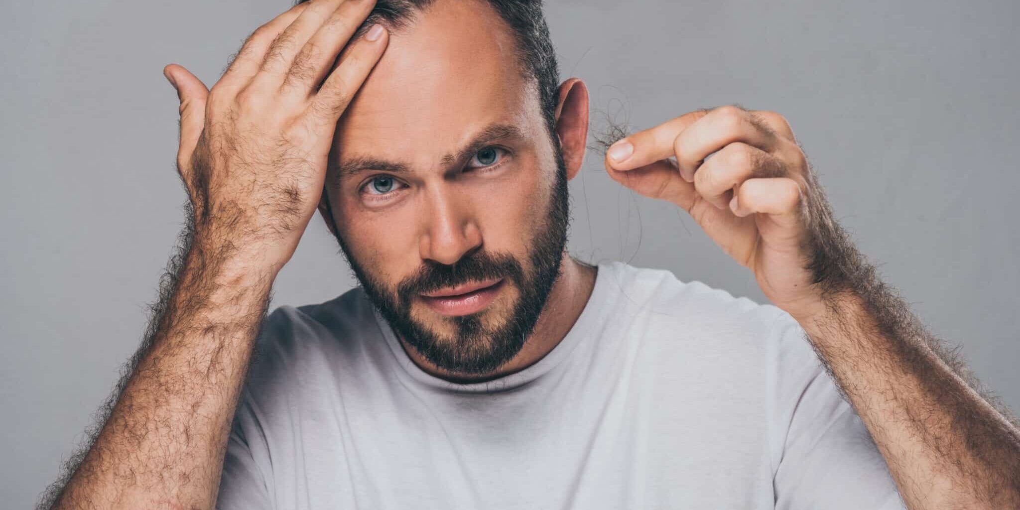 bearded middle aged man holding fallen hair and looking at camera isolated on grey prp behandling
